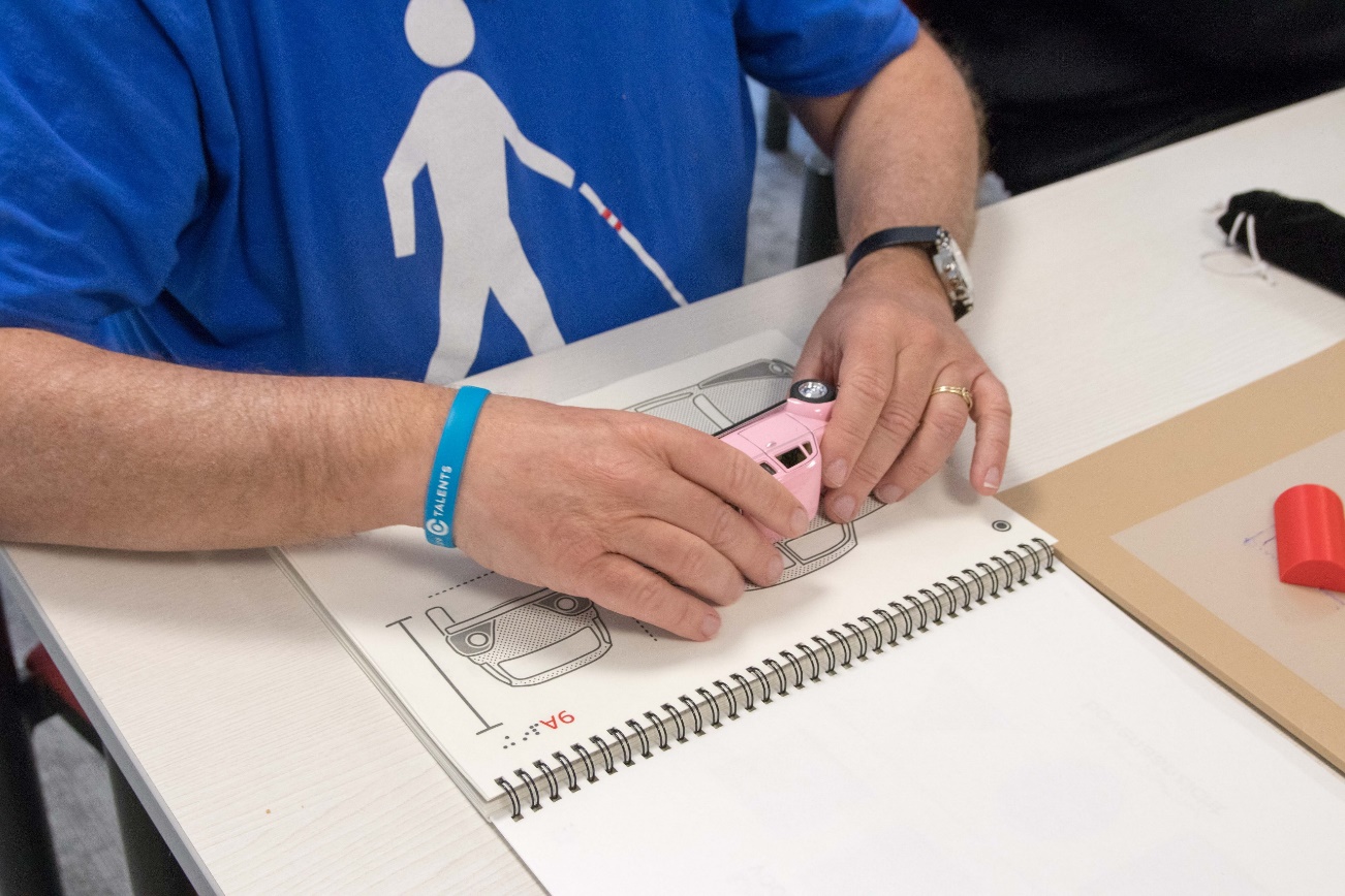 Iemand heeft een speelgoedauto vast en daaronder een pagina met de voelbare tekening van een auto.