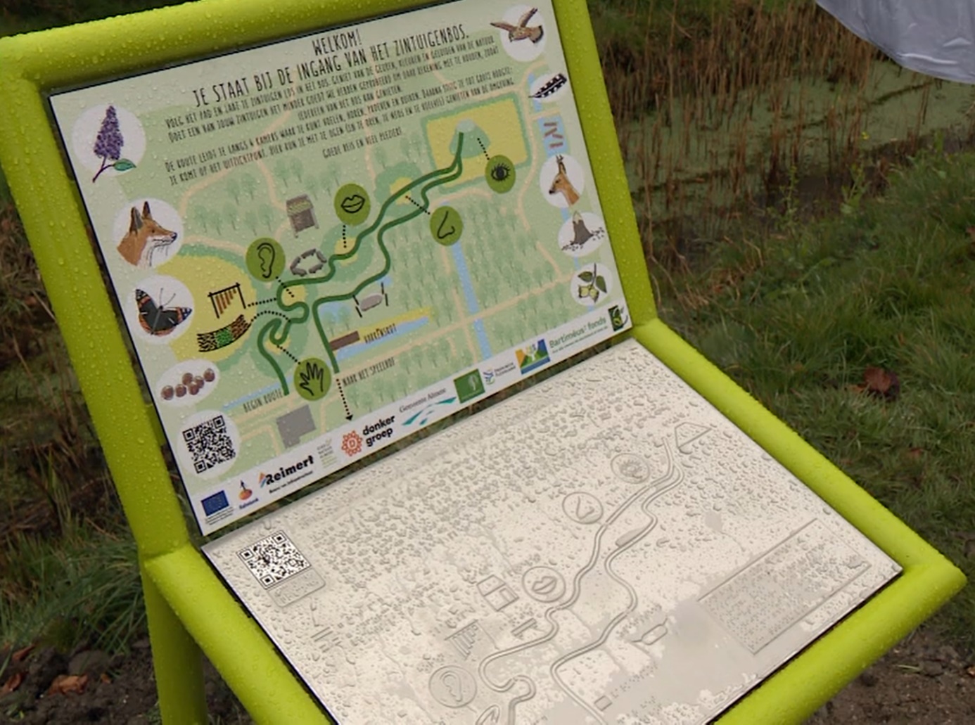 Een informatiebord met de wandelroute van het Zintuigenbos in Almere. Bovenaan staat het in kleur en grootletter, onderaan een metalen plaat met tekst in braille en reliëf